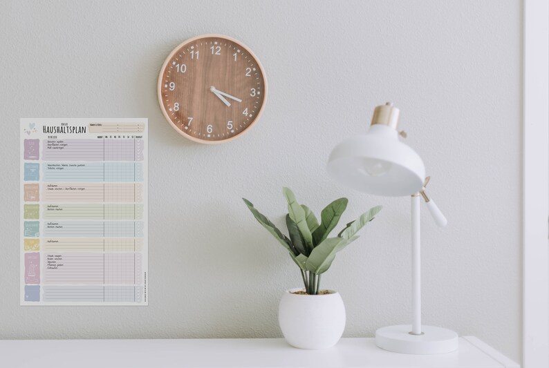 May include: A white desk with a wooden clock, a white lamp, and a potted plant. A printed sheet with the text "Haushaltsplan" is on the wall.
