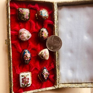 May include: A collection of small, decorative cloisonn&eacute; boxes in various shapes, including square, oval, and diamond. Each box features floral designs in shades of pink, blue, and green, set against a black, white, or blue background. The boxes are displayed in a red velvet-lined case.