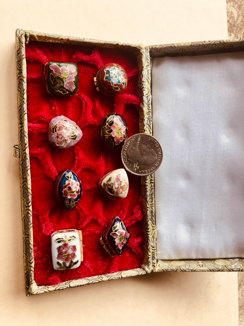 May include: A collection of small, decorative cloisonn&eacute; boxes in various shapes, including square, oval, and diamond. Each box features floral designs in shades of pink, blue, and green, set against a black, white, or blue background. The boxes are displayed in a red velvet-lined case.