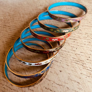 May include: A set of six colorful enamel bangles with floral designs. The bangles are stacked on top of each other, with the turquoise bangle on top. The bangles are decorated with gold trim.