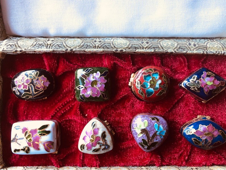 May include: A collection of eight small, decorative cloisonn&eacute; boxes in various shapes, including oval, square, and heart. Each box features a floral design with pink, blue, and gold accents, set against a black, red, green, white, and blue background. The boxes are displayed in a red velvet-lined case.