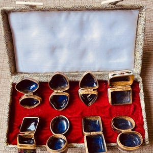 May include: An open box with a red velvet interior displays several small, hinged, gold-toned trinket boxes. The boxes have various shapes, including oval, heart, and square, with deep blue enamel interiors. The box itself has a patterned exterior.