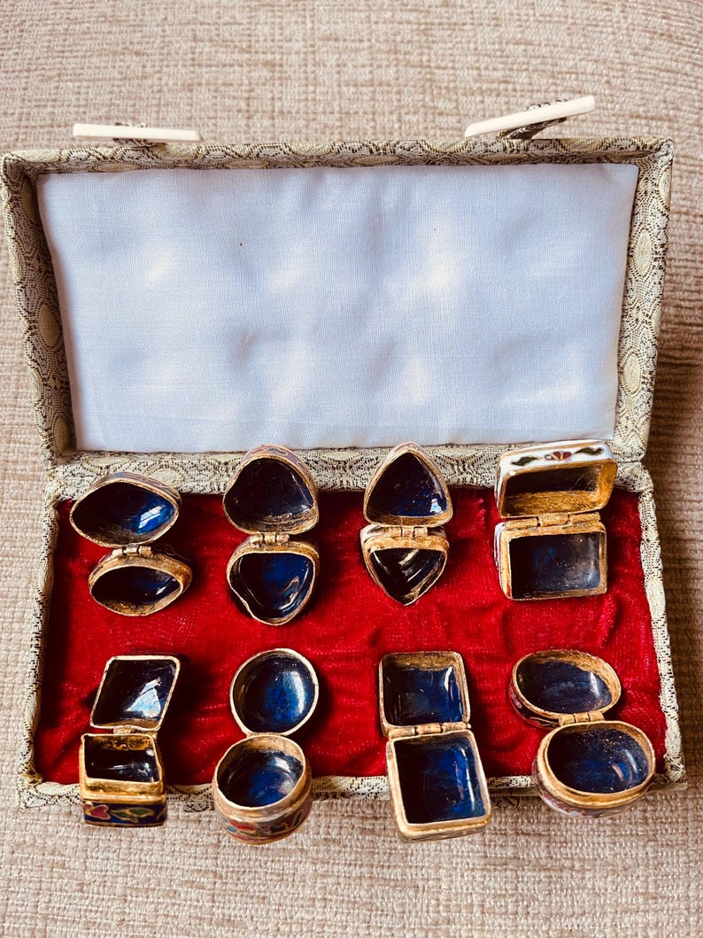 May include: An open box with a red velvet interior displays several small, hinged, gold-toned trinket boxes. The boxes have various shapes, including oval, heart, and square, with deep blue enamel interiors. The box itself has a patterned exterior.