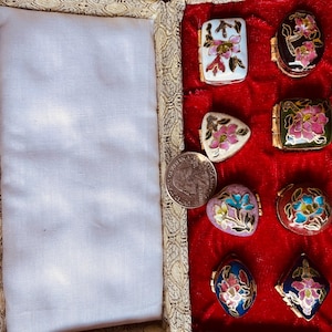 May include: A collection of small, decorative trinket boxes with floral designs in various shapes, including rectangular, oval, and triangular. The boxes are set against a red velvet background within a patterned case. A US quarter is also visible.