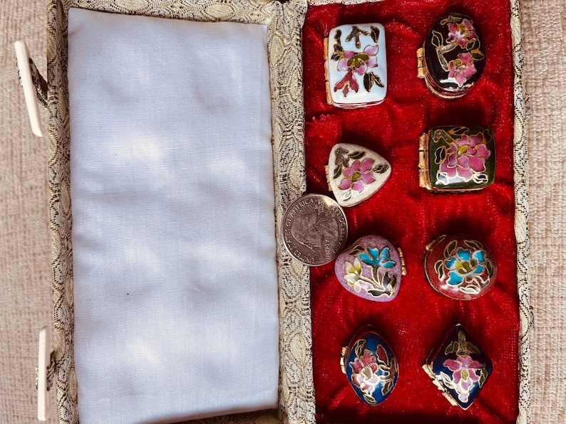 May include: A collection of small, decorative trinket boxes with floral designs in various shapes, including rectangular, oval, and triangular. The boxes are set against a red velvet background within a patterned case. A US quarter is also visible.