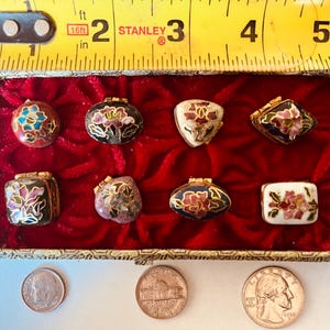 May include: A collection of eight small, decorative trinket boxes with floral designs in a variety of shapes. The boxes are displayed in a red velvet-lined box. A ruler shows the box is approximately 3 inches wide.