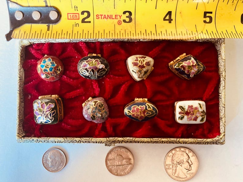 May include: A collection of eight small, decorative trinket boxes with floral designs in a variety of shapes. The boxes are displayed in a red velvet-lined box. A ruler shows the box is approximately 3 inches wide.