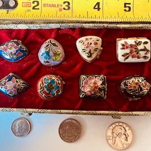 May include: A collection of eight small, decorative trinket boxes in various shapes, including oval, square, and triangular, with floral designs. The boxes are displayed in a red velvet-lined box. A measuring tape shows the box is approximately 6 inches long.