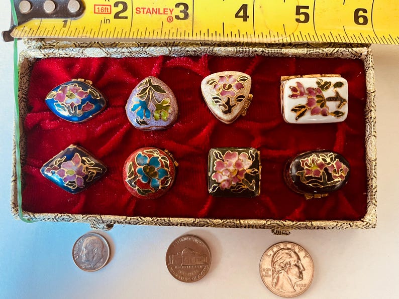 May include: A collection of eight small, decorative trinket boxes in various shapes, including oval, square, and triangular, with floral designs. The boxes are displayed in a red velvet-lined box. A measuring tape shows the box is approximately 6 inches long.