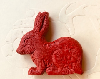 Peut inclure: Une figurine de lapin rouge sculptée avec des motifs floraux détaillés. Le lapin est assis sur une surface blanche.