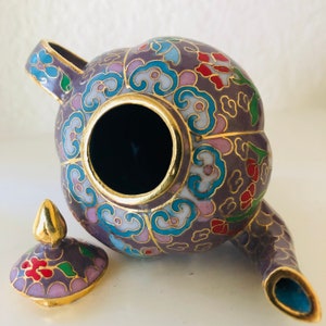 Può includere: Una piccola teiera in cloisonné viola con un coperchio e motivi floreali. La teiera è in metallo e ha un bordo dorato.
