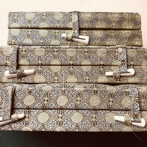 May include: Three stacked decorative boxes with a repeating geometric pattern in shades of gray, beige, and brown. Each box has a fabric strap closure with a white toggle. The boxes are likely for storage or display.