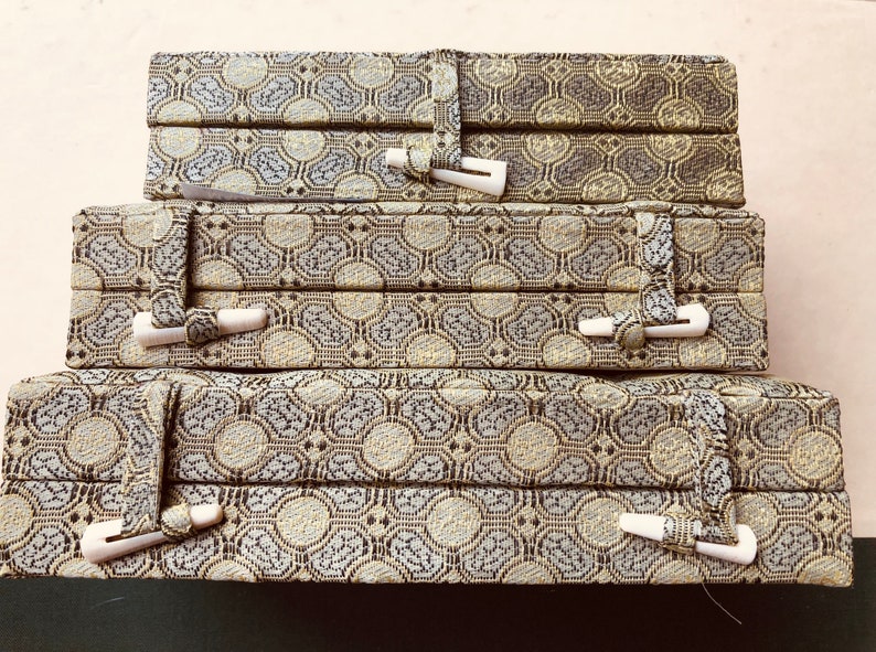 May include: Three stacked decorative boxes with a repeating geometric pattern in shades of gray, beige, and brown. Each box has a fabric strap closure with a white toggle. The boxes are likely for storage or display.