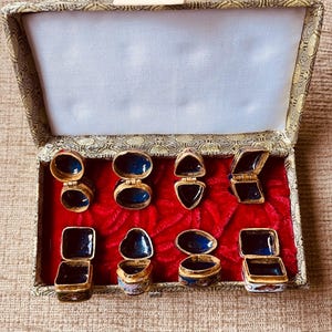 May include: A collection of small, ornate trinket boxes displayed in an open, fabric-lined box. The boxes are gold-toned with blue enamel interiors and lids. The open box reveals a red velvet base and a cream-colored interior.