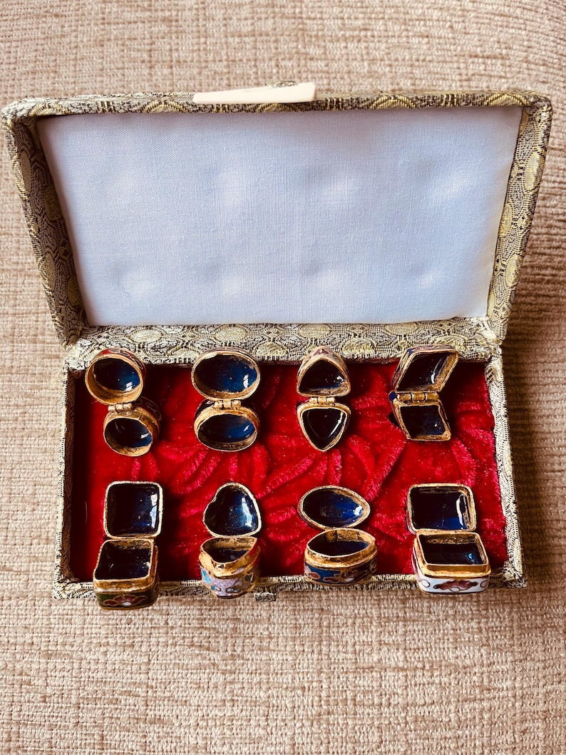 May include: A collection of small, ornate trinket boxes displayed in an open, fabric-lined box. The boxes are gold-toned with blue enamel interiors and lids. The open box reveals a red velvet base and a cream-colored interior.