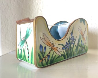 May include: A white ceramic tape dispenser with a gold trim and a dragonfly design. The dispenser has a blue and green floral pattern and is decorated with dragonflies.