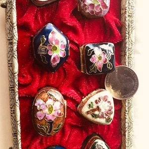 May include: A collection of small, decorative enamel boxes in various shapes, including oval, square, and heart-shaped. Each box features floral designs in pink, white, and blue, set against a red velvet background.