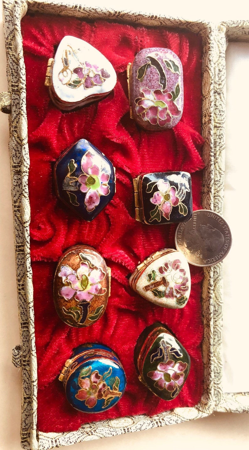 May include: A collection of small, decorative enamel boxes in various shapes, including oval, square, and heart-shaped. Each box features floral designs in pink, white, and blue, set against a red velvet background.
