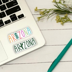 May include: A white laptop computer with a sticker that says "ARIZONA" in rainbow letters with a pine tree and a cactus graphic. The laptop is on a white wooden surface with a green pen and a bouquet of white flowers.