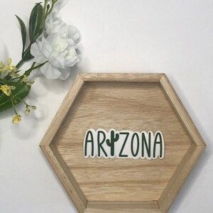 May include: A green and white sticker with the word "ARIZONA" and a cactus graphic on a wooden hexagonal tray.
