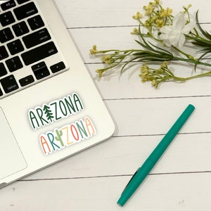 May include: A laptop computer with two stickers on it that say "Arizona" in different fonts and colors. The stickers are on a white background. A green pen is on a white wooden surface.