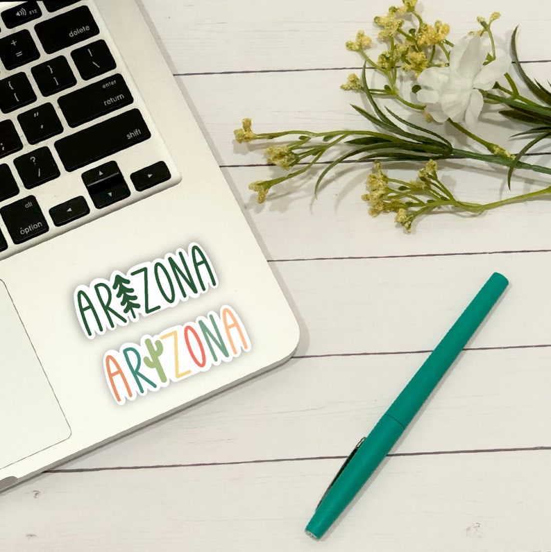 May include: A laptop computer with two stickers on it that say "Arizona" in different fonts and colors. The stickers are on a white background. A green pen is on a white wooden surface.