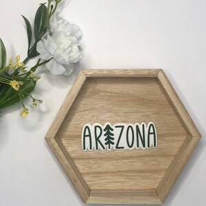 May include: A green and white sticker with the word "ARIZONA" and a stylized pine tree in the middle of the word. The sticker is on a wooden hexagonal tray.