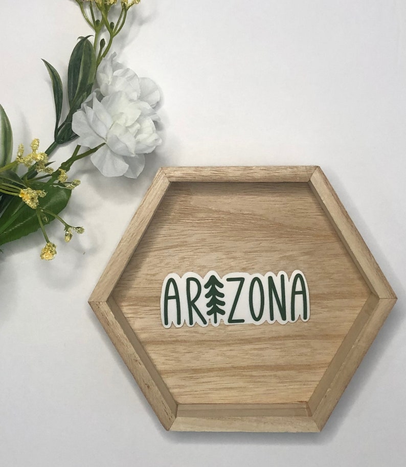 May include: A green and white sticker with the word "ARIZONA" and a stylized pine tree in the middle of the word. The sticker is on a wooden hexagonal tray.