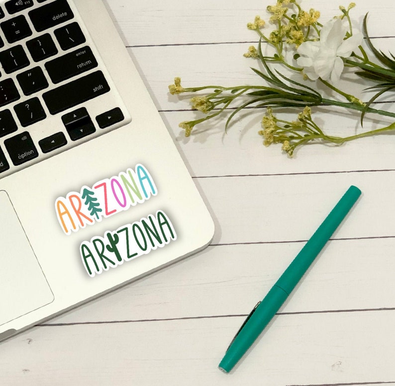 May include: A white laptop computer with a sticker that says "ARIZONA" in rainbow letters with a pine tree and a cactus graphic. The laptop is on a white wooden surface with a green pen and a bouquet of white flowers.