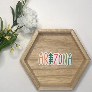 May include: A colorful sticker with the word "ARIZONA" in rainbow colors with a green pine tree in the middle of the word. The sticker is on a hexagonal wooden tray.