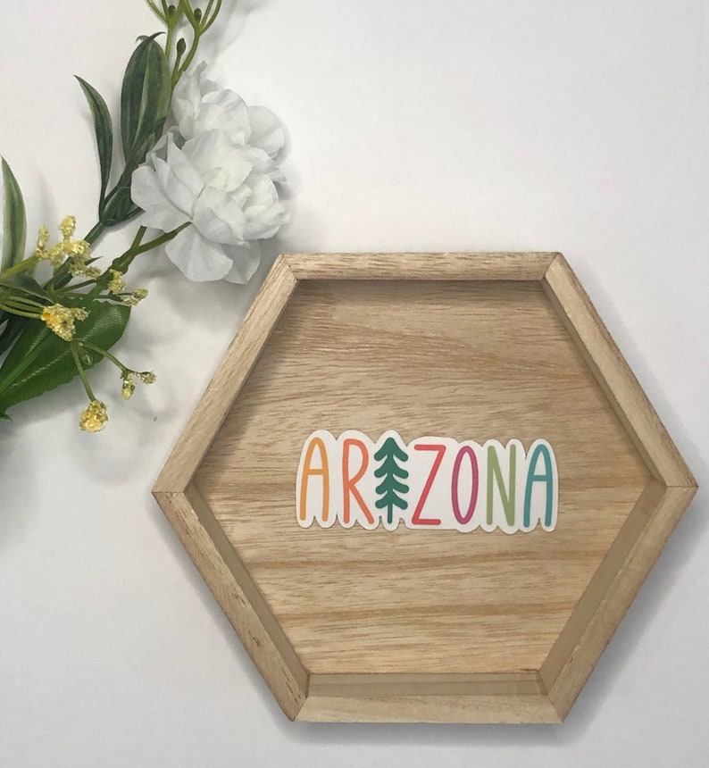 May include: A colorful sticker with the word "ARIZONA" in rainbow colors with a green pine tree in the middle of the word. The sticker is on a hexagonal wooden tray.