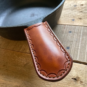May include: A brown leather pot holder with decorative stitching around the edges. The holder is attached to a cast iron skillet.