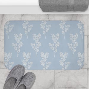 Könnte beinhalten: Eine rechteckige, hellblaue Badematte mit einem sich wiederholenden Muster aus weißen Blumenzweigen. Die Matte liegt auf einem gefliesten Boden, mit einem Paar grauer Hausschuhe und einem grauen Handtuch.