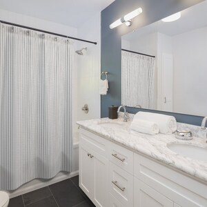 May include: A contemporary bathroom featuring a white vanity unit with a marble countertop and a large mirror. A striped shower curtain is visible. The walls are painted white and a dark blue-grey. Two white towels are neatly folded on the counter.