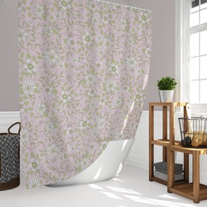 May include: A pink shower curtain with a floral pattern featuring white flowers and green leaves. The shower curtain is hanging in a bathroom with a white bathtub and a wooden shelf with towels and a basket.