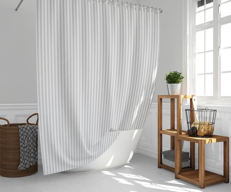 May include: A grey and white striped shower curtain in a bathroom setting. A brown wicker basket with a grey blanket is on the floor. Wooden shelves display a potted plant and other bathroom accessories. A large window allows natural light.