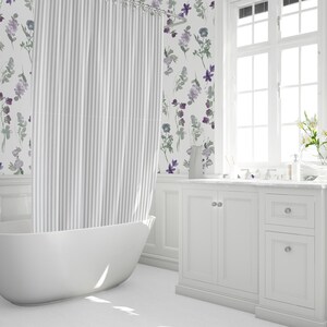 May include: A bright, white bathroom with a freestanding tub and a striped shower curtain. The walls feature floral wallpaper in shades of purple and green. A white vanity with a marble countertop and a large window complete the space.