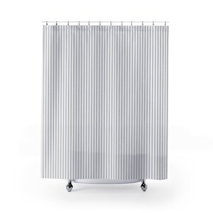 May include: A grey and white striped shower curtain suspended from a metal rail with silver hooks. The curtain features vertical stripes of varying widths, offering a classic, clean aesthetic. The curtain hangs in front of a white shower base.