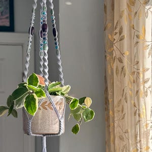 May include: A hanging macrame plant holder in light gray, adorned with beads in shades of purple, blue, and green. The holder suspends a ceramic pot with a plant featuring green and white leaves. The background includes a window with patterned curtains.