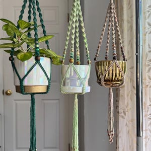 May include: Three macrame plant hangers suspended from wooden rings. The left hanger is dark green with a white iridescent pot. The center hanger is light green with a white iridescent pot. The right hanger is beige with a metallic gold fluted pot.