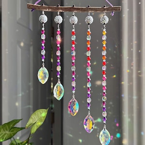 Puede incluir: Móvil decorativo colgante con una rama de madera natural, cinco hilos de cuentas de colores y colgantes de cristal iridiscente. Cada hilo tiene una bola plateada en la parte superior. Las cuentas son en tonos morado, rosa, rojo, naranja y amarillo.