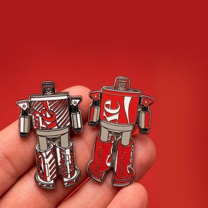 May include: Two robot-shaped enamel pins. One pin features a red and white design with the text "Re" and the other pin features a red and white design with the text "Re". The pins have a silver-colored body and legs.