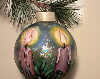 Könnte beinhalten: Ein handbemalter Weihnachtsbaumschmuck aus Glas mit zwei lila Kerzen mit gelben Flammen vor einem blauen und grünen Hintergrund mit Glitzer.