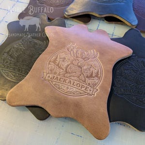 May include: Leather coasters in various colors, featuring a detailed embossed design of a jackalope within a circular frame, with the word "JACKALOPE" across a banner. The coasters are from Noble Buffalo Handmade Leather Goods.