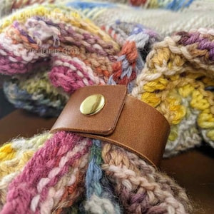Könnte beinhalten: Ein braunes Lederband mit einer goldenen Metallschnalle, umwickelt um einen bunten Häkel-Schal. Der Schal hat eine Vielzahl von Farben, darunter Pink, Gelb, Blau und Grün. Das Band ist schlicht gehalten und hat eine glatte Lederoberfläche. Das Band ist eine großartige Möglichkeit, einen Schal an Ort und Stelle zu halten und einen Hauch von Stil hinzuzufügen.