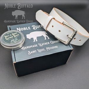 May include: A white leather belt with a silver buckle rests on a gray box. The box is labeled "Noble Buffalo Handmade Leather Goods" and includes a tin of "Belt & Boot Leather Balm". The text "Saint Louis, Missouri" is also visible.
