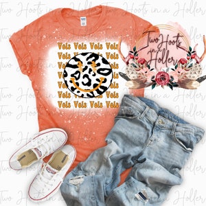 Puede incluir: Una camiseta naranja con un diseño salpicado de lejía que presenta una cara sonriente de estampado de leopardo blanco y negro y el texto "Vols Vols Vols Vols" repetido. La camiseta se combina con unos vaqueros azules y zapatillas blancas. También se incluye un gráfico rosa y dorado con el texto "Two Hoots in a Holler".