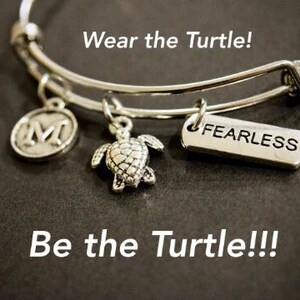 May include: Silver charm bracelet with a turtle, letter M, and "Fearless" charm. The bracelet is on a dark background with the text "Wear the Turtle!" and "Be the Turtle!!!"