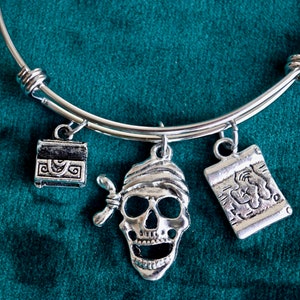 May include: Silver-toned expandable bracelet with three pirate-themed charms. Charms include a treasure chest, a skull wearing a bandana, and a scroll. The bracelet is displayed on a dark teal background.