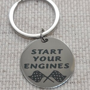 May include: Silver keychain with a round metal tag that says "Start Your Engines" with two black and white checkered flags crossed behind the text.